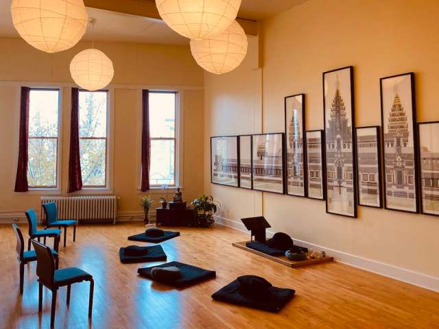 Photograph of the sitting space in the Northfield Buddhist Meditation Center.  Chairs and cushions facing a wall with multi-paneled artwork representing a temple.